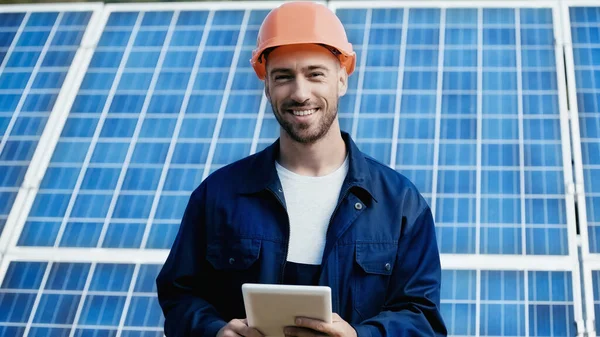 Happy engineer in hardhat holding gadget near solar panel — Stock Photo
