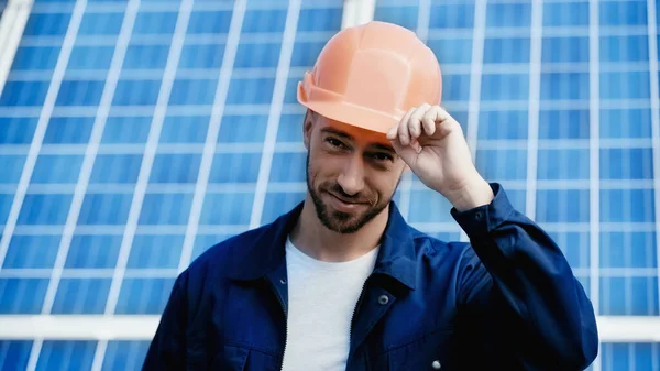 Pleased engineer looking at camera and adjusting orange helmet — Foto stock