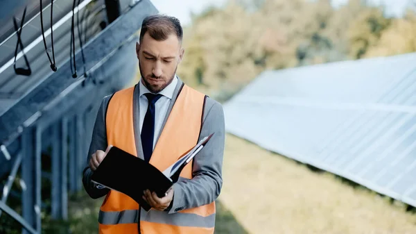 Businessman in safety vest holding folder with documents near solar panels — Stockfoto