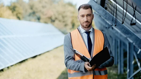 Businessman in safety vest and suit holding folder near solar panels — Foto stock
