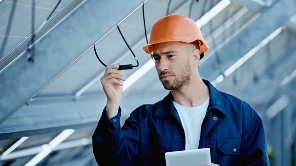 Engineer in hardhat checking wire of solar panels while holding digital tablet - foto de stock