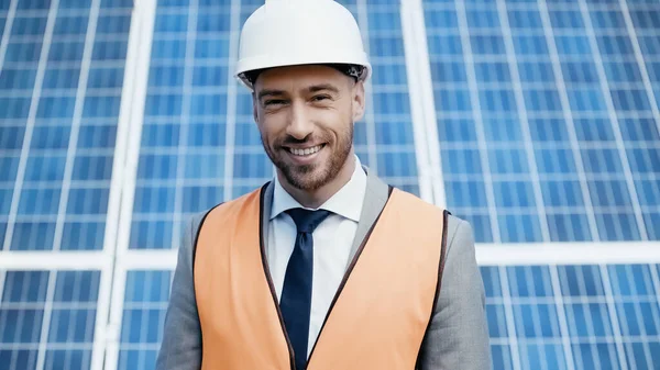 Cheerful businessman in hardhat and safety vest near solar batteries — Foto stock