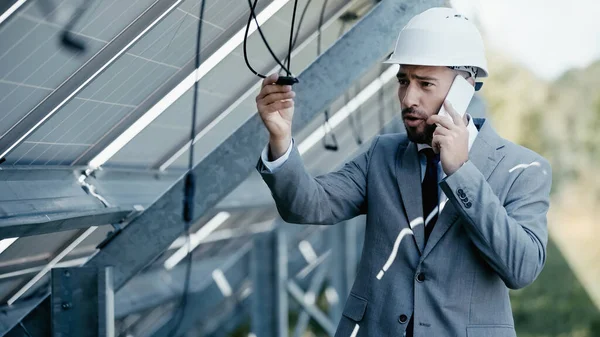 Dissatisfied businessman looking at hanging wire while talking on smartphone near solar panels — Stock Photo