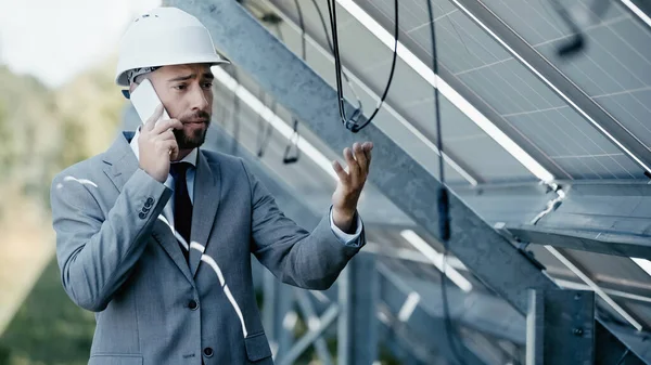 Verärgerter Geschäftsmann schaut auf hängenden Draht, während er in der Nähe von Sonnenkollektoren mit dem Smartphone spricht — Stockfoto