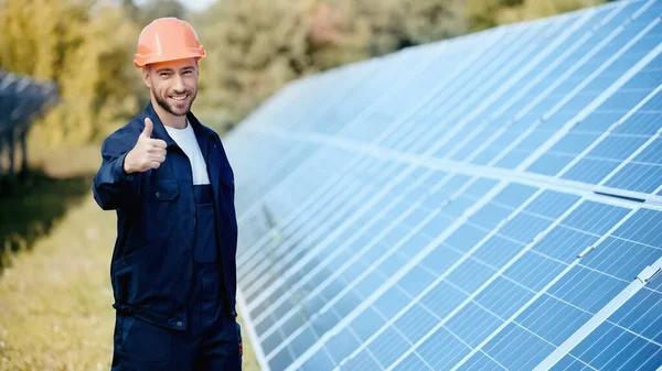 Happy engineer in hardhat showing thumb up near solar batteries — Stock Photo