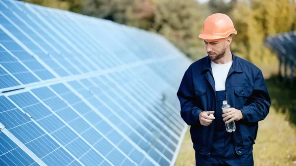 Engineer in hardhat and safety vest holding bottle with water near solar batteries — Stock Photo