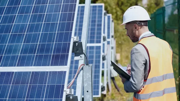 Businessman in hardhat looking at wires and holding folder near solar batteries outside — Stockfoto