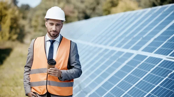 Smiling businessman in hardhat holding takeaway beverage near solar batteries outside — Stock Photo