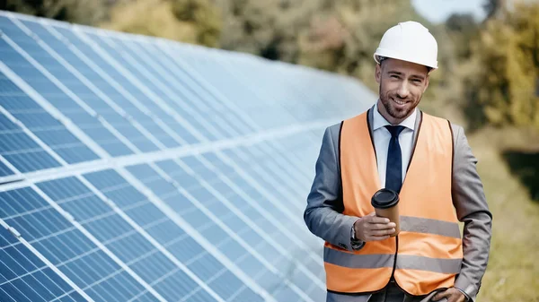 Glücklicher Geschäftsmann in Hardhat hält Imbissgetränk in der Nähe von Solarbatterien draußen — Stockfoto