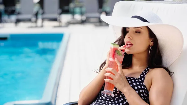 Bruna giovane donna in costume da bagno e cappello di paglia bere cocktail mentre sdraiato sul lettino — Foto stock