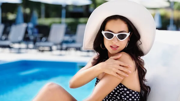 Brunette woman with straw hat and sunglasses applying sunscreen on arm near swimming pool — Stock Photo