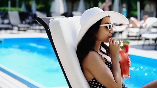 Vista lateral de mujer morena con sombrero de sol y gafas de sol bebiendo cóctel de paja - foto de stock