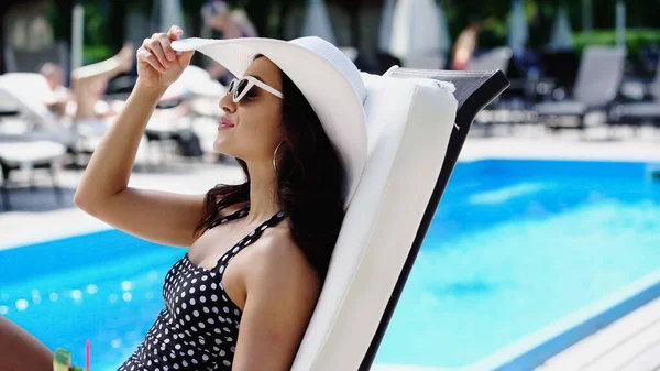 Brunette woman in sunglasses adjusting straw hat while lying on lounger — Stock Photo