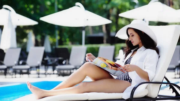Barefoot woman in straw hat reading magazine while resting on lounger near swimming pool — Stock Photo