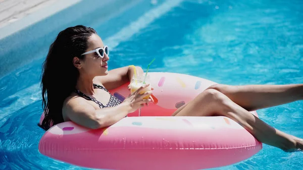 Happy woman in sunglasses holding glass with cocktail and swimming on inflatable ring in pool — Stock Photo