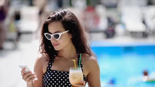 Mujer alegre en gafas de sol y traje de baño celebración de cóctel y el uso de teléfono inteligente - foto de stock