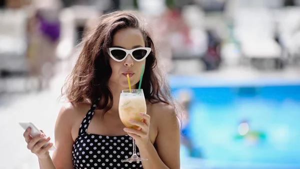 Brunette woman in sunglasses and swimsuit drinking cocktail and holding smartphone - foto de stock
