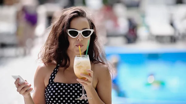 Brunette woman in sunglasses and swimsuit drinking cocktail and holding smartphone — Stock Photo