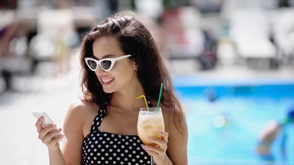 Happy woman in sunglasses and swimsuit holding cocktail and using smartphone — Stock Photo
