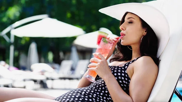 Brunette young woman in dotted swimsuit and straw hat drinking cocktail while lying on lounger — Stockfoto