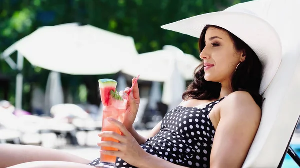 Happy young woman in dotted swimsuit and straw hat holding cocktail while lying on lounger — Stock Photo