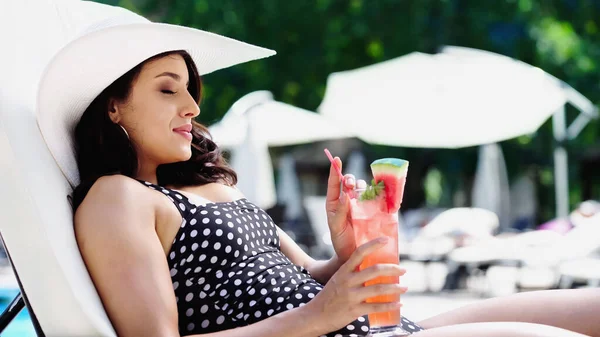 Pleased woman in dotted swimsuit and straw hat drinking cocktail while lying on lounger — Stock Photo