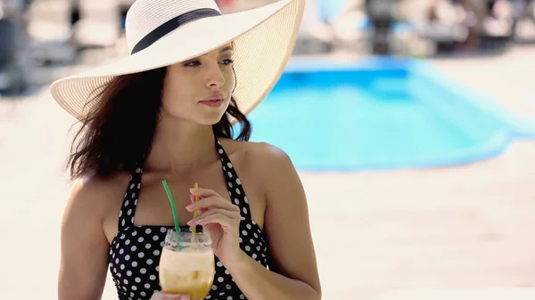 Brunette young woman in dotted swimsuit and straw hat holding glass with cocktail — Stockfoto