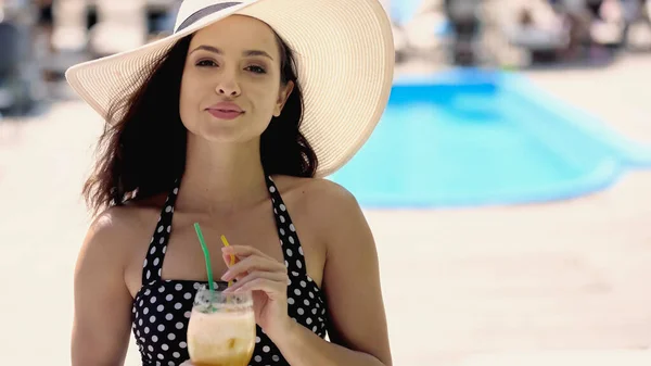 Pleased young woman in dotted swimsuit and straw hat holding glass with cocktail — Stockfoto