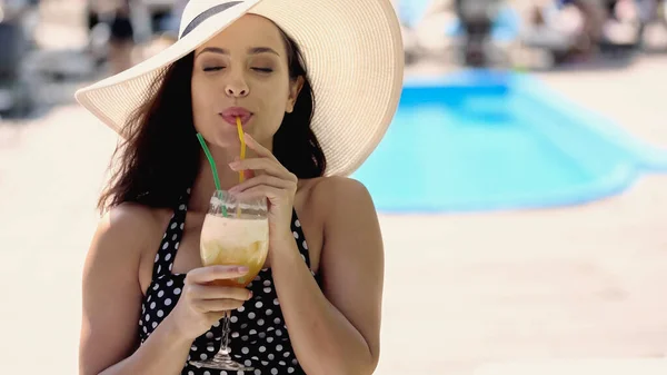 Brunette young woman in dotted swimsuit and straw hat drinking refreshing cocktail — Stockfoto