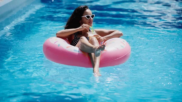 Happy woman in stylish sunglasses holding glass with cocktail and swimming on inflatable ring in pool — Stock Photo