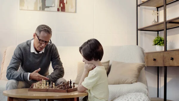Avô em óculos e neto inteligente jogando xadrez na sala de estar — Fotografia de Stock