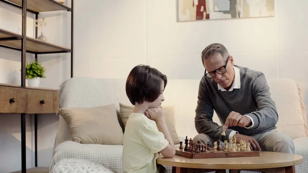 Grandfather in glasses and preteen grandson playing chess in living room — стоковое фото