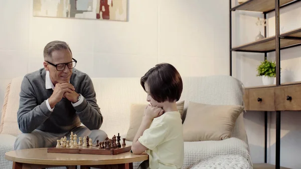 Happy grandfather in glasses and smart grandson playing chess in living room — Foto stock