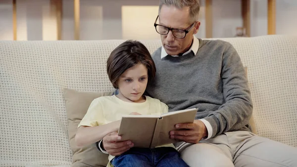 Grandfather in glasses reading book with grandson at home — Stockfoto