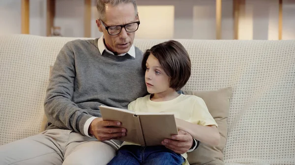 Grandfather in glasses reading book to grandson at home - foto de stock