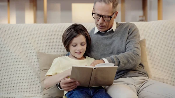 Grandfather in glasses reading book with happy grandson at home — Stock Photo