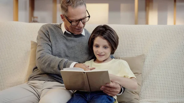 Cheerful grandfather in glasses reading book with grandson at home — Stock Photo