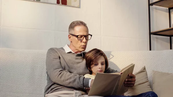 Grandfather in glasses reading book to preteen grandson at home — Stock Photo