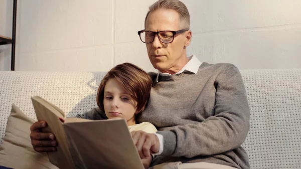 Mature grandfather in glasses reading book with grandson at home — Stockfoto