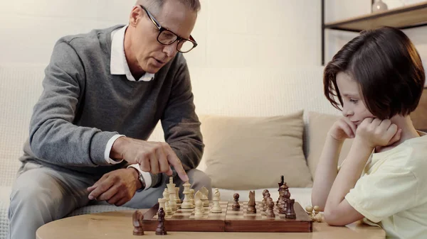 Grand-père dans les lunettes pointant vers la figure d'échecs dans le salon — Photo de stock