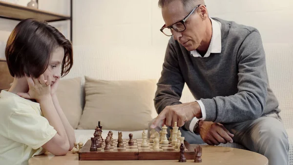 Grand-père dans des lunettes pointant vers la figure près du petit-fils blanc jouer aux échecs dans le salon — Photo de stock