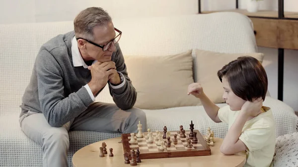Grand-père dans des lunettes regardant petit-fils intelligent tout en jouant aux échecs dans le salon — Photo de stock