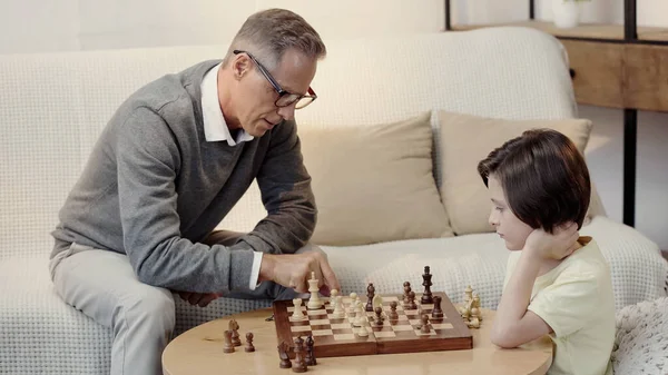 Abuelo en gafas y preadolescente niño jugando ajedrez en sala de estar - foto de stock