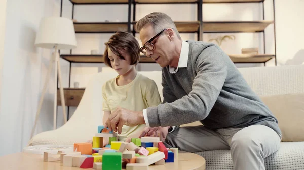 Abuelo de mediana edad en gafas jugando bloques de construcción con nieto preadolescente - foto de stock