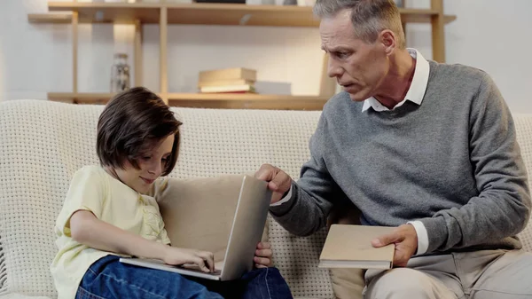 Grand-père d'âge moyen tenant livre près de petit-enfant à l'aide d'un ordinateur portable — Photo de stock
