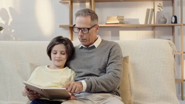 Middle aged grandfather in glasses reading book with grandson at home — Stock Photo