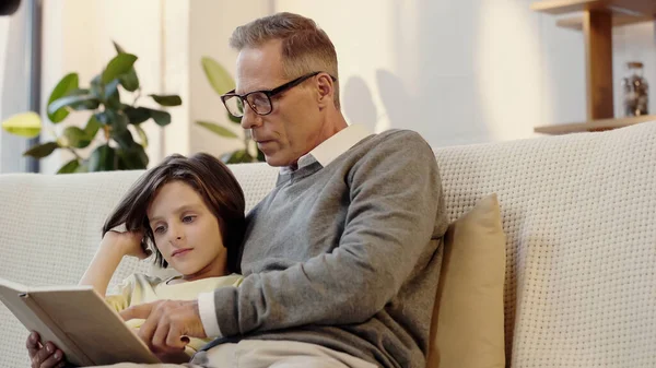 Middle aged grandfather in glasses reading book with boy at home — Stock Photo