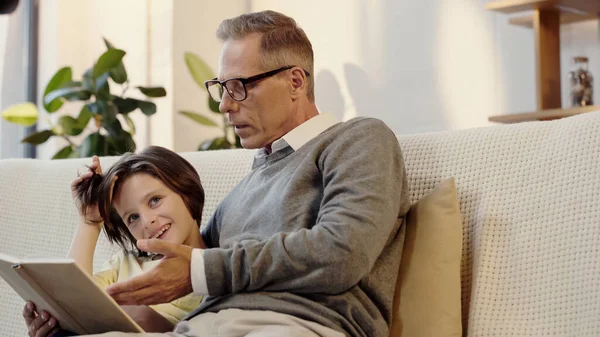 Middle aged grandfather in glasses reading book with cheerful grandson at home - foto de stock