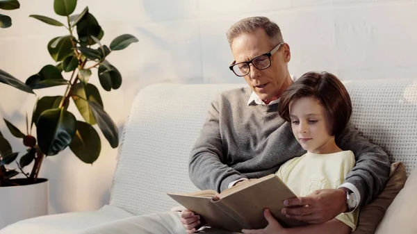 Middle aged grandfather in glasses reading book to kid at home — Stock Photo