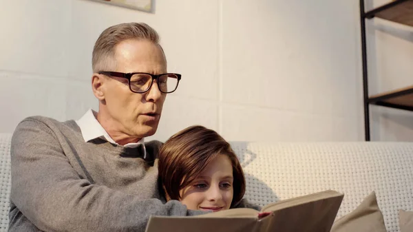 Großvater mittleren Alters mit Brille liest Buch mit lächelndem Enkel zu Hause — Stockfoto